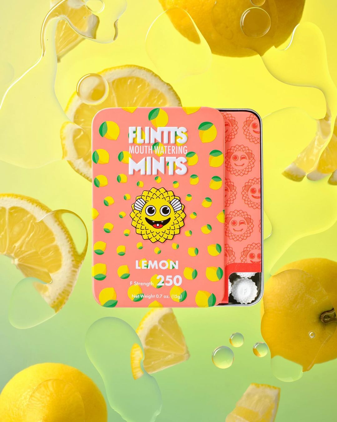 Eye-level shot of a pink tin of Flintt's Lemon Mouth Watering Mints floating amongst slices of lemons and lemon juice on a yellow to green gradient background.