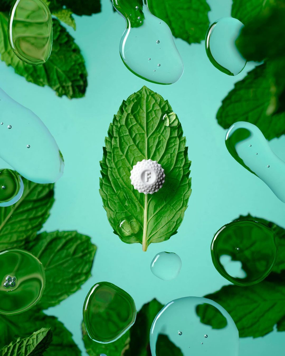 Mint leaf with a small white circular object on top, surrounded by water droplets on a light blue background, food photography.