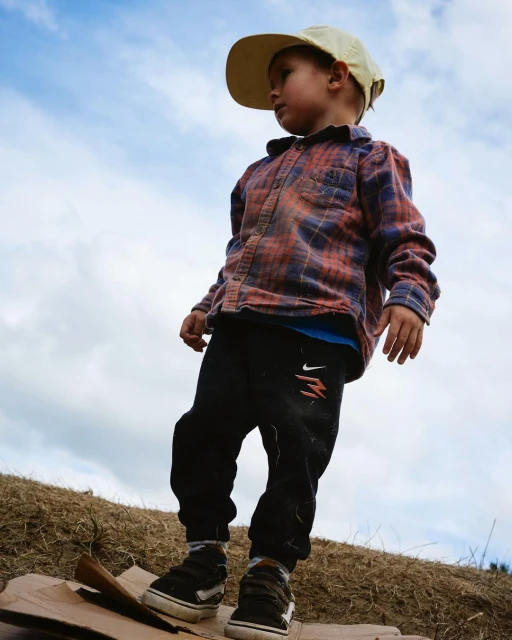 Low-angle view of a toddler in a flannel shirt and baseball cap standing on cardboard outdoors with a cloudy sky in the background.