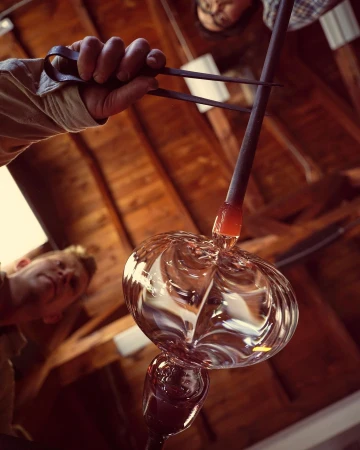 Craftsman shaping molten glass with tools in a workshop, creating a translucent glass form with wood beams and rafters in background.