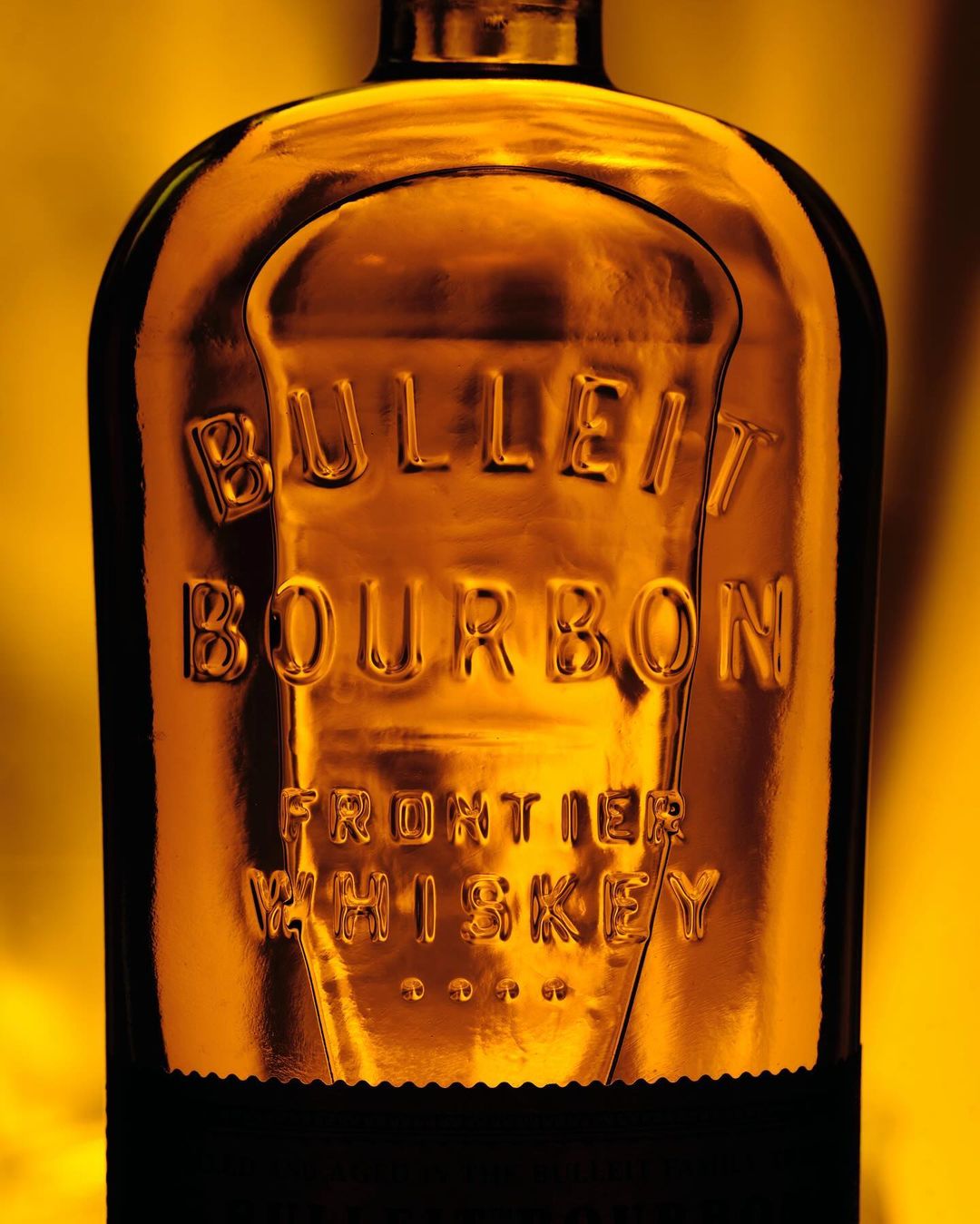 Close-up of a Bulleit Bourbon bottle with embossed lettering, showcasing the amber liquid inside against a blurred yellow background.