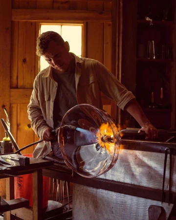 Adult male shaping molten glass with a blowtorch in a workshop, crafting a clear, round vase against a warm, rustic interior.