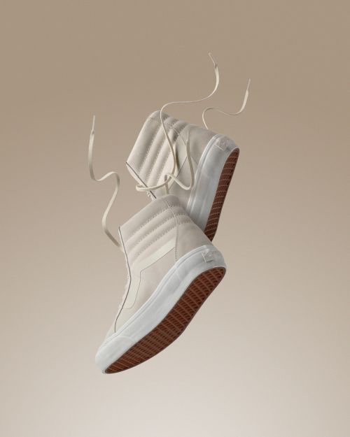 Pair of high-top Vans sneakers in light beige, angled with laces floating, against a soft beige background. Studio product shot for fashion.