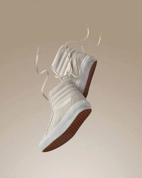 Pair of high-top Vans sneakers in light beige, angled with laces floating, against a soft beige background. Studio product shot for fashion.