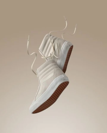 Pair of high-top Vans sneakers in light beige, angled with laces floating, against a soft beige background. Studio product shot for fashion.