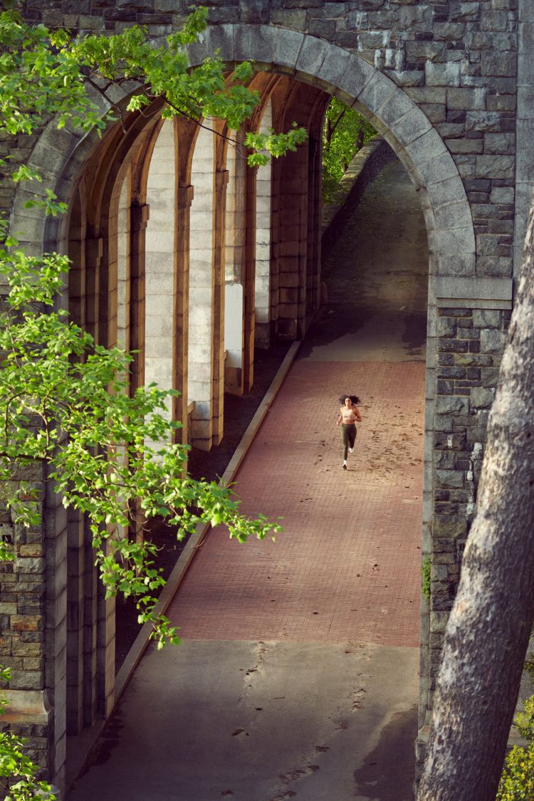 A woman jogs on a brick path underneath stone arches surrounded by lush greenery, viewed from a high angle, highlighting fitness and architectural beauty.