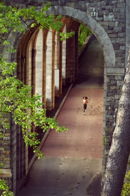 A woman jogs on a brick path underneath stone arches surrounded by lush greenery, viewed from a high angle, highlighting fitness and architectural beauty.