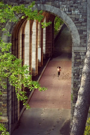 A woman jogs on a brick path underneath stone arches surrounded by lush greenery, viewed from a high angle, highlighting fitness and architectural beauty.