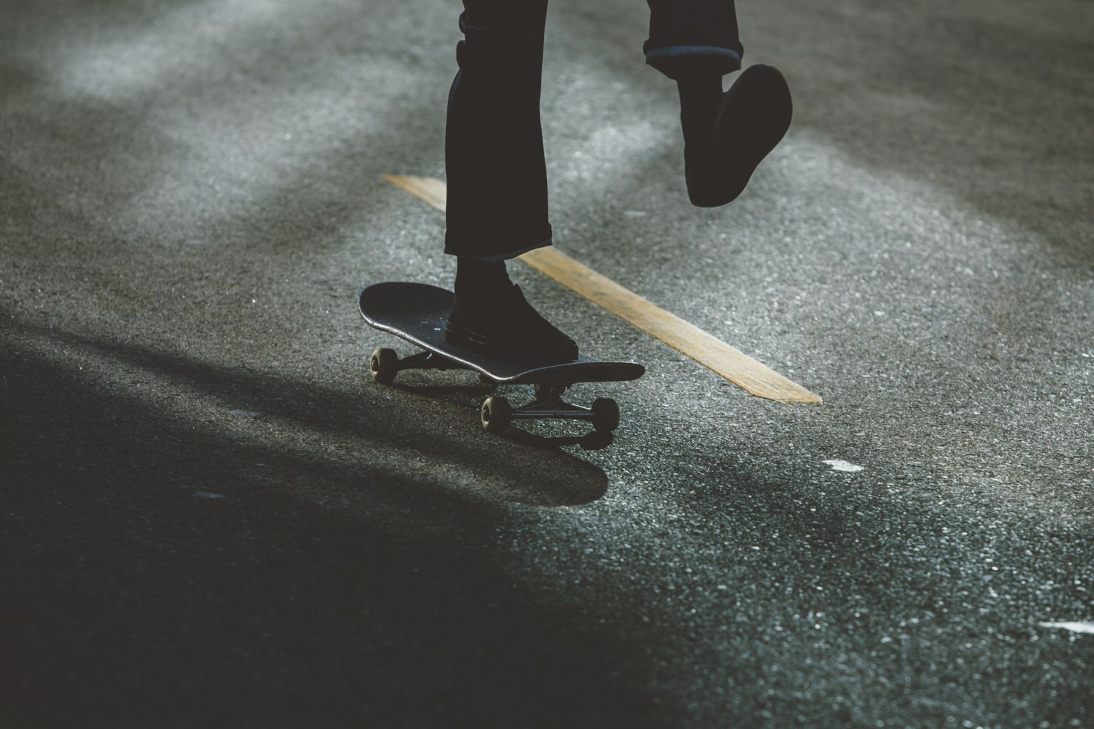 Person skateboarding on asphalt, captured from a low angle with dark color grading, focusing on movement and texture in an outdoor environment.