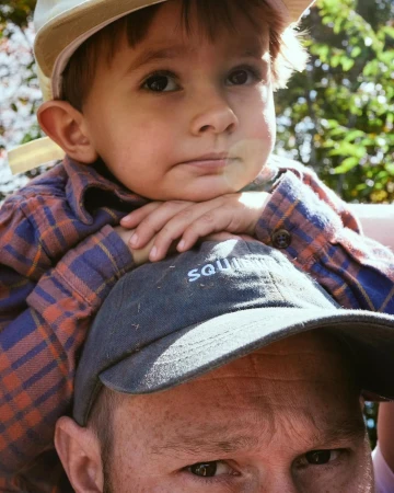 A young child sitting on a man's shoulders, wearing a plaid shirt and light cap, photographed in Los Angeles by Brian Kelley.