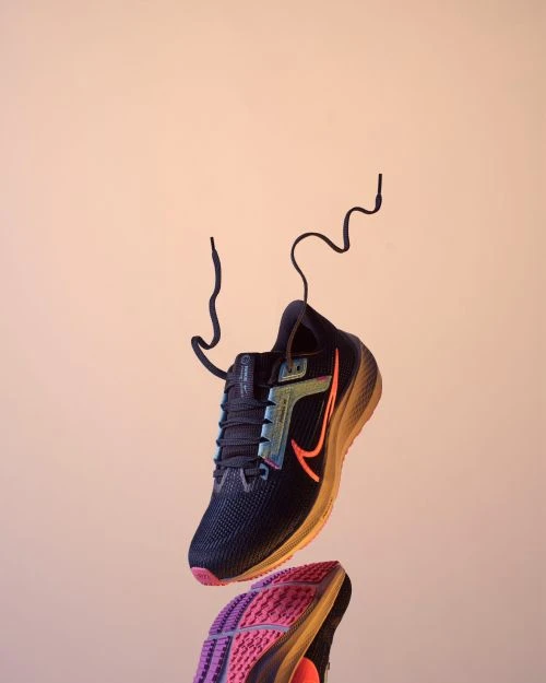 Athletic shoe suspended in studio shot, angled view showing black mesh upper, pink and purple sole, orange Nike swoosh logo detail.