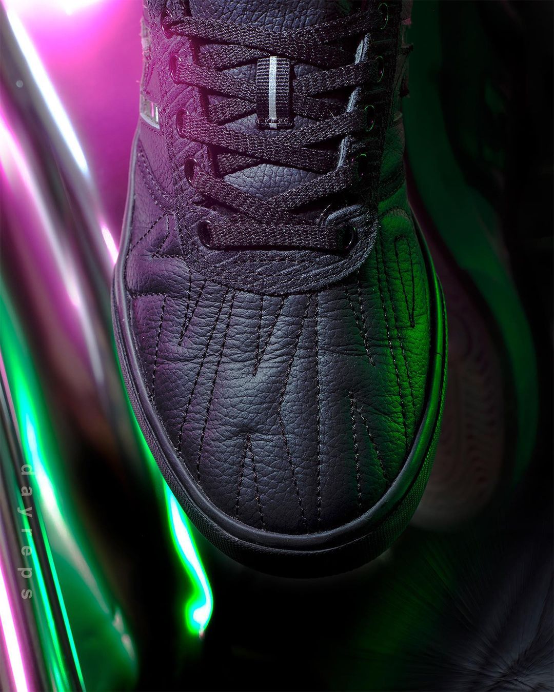 Close-up of a black sneaker with laces against an abstract background with pink and green light reflecting off metallic material.