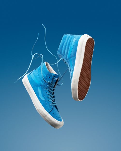 Studio shot of two floating, bright blue, high-top Vans sneakers with white soles against a gradient blue background.