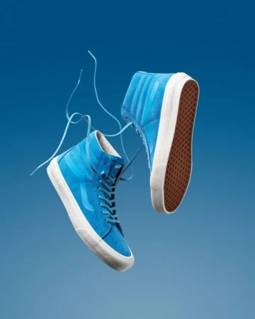 Studio shot of two floating, bright blue, high-top Vans sneakers with white soles against a gradient blue background.
