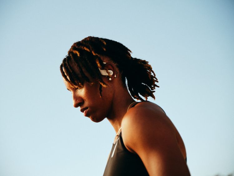 Close-up of an adult woman with locs, wearing wireless earbuds and a black tank top, set against a clear blue sky.