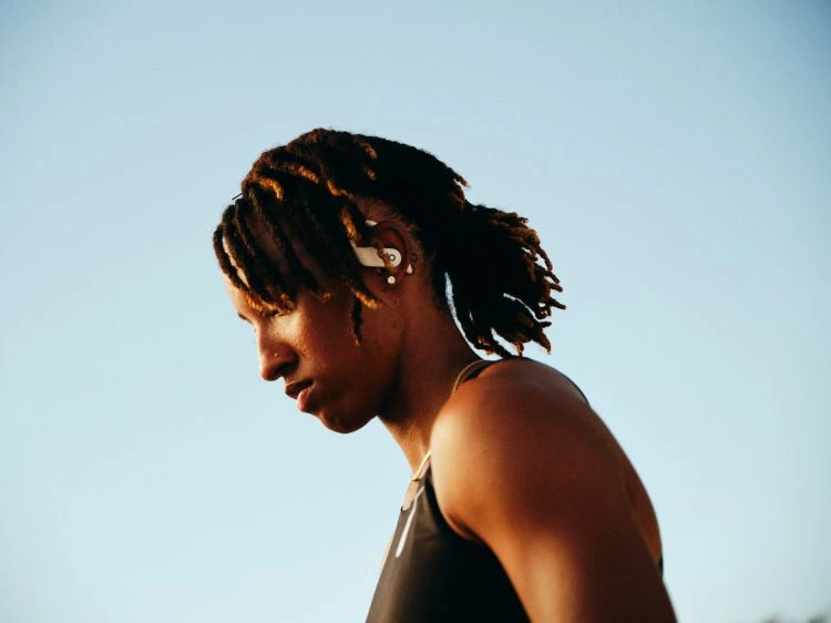 Close-up of an adult woman with locs, wearing wireless earbuds and a black tank top, set against a clear blue sky.