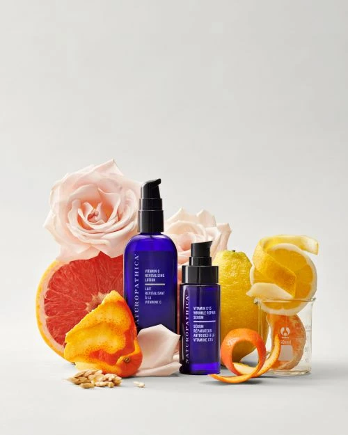 Two Naturopathica skincare bottles surrounded by roses and various citrus fruits with peels on a white surface in a studio setting.
