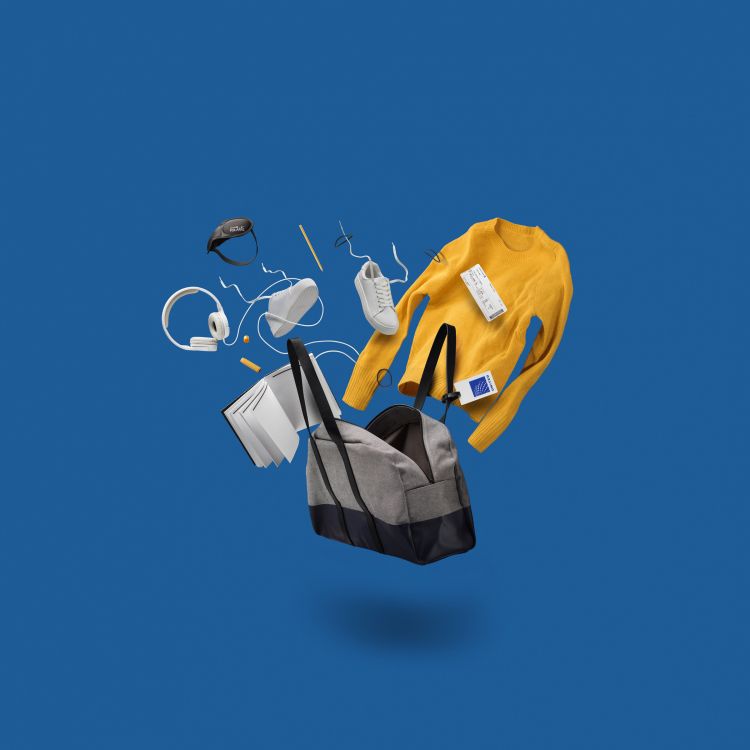 Elevated view of a gray duffel bag surrounded by a yellow sweater, white shoes, headphones, a book, and other objects against a blue backdrop.