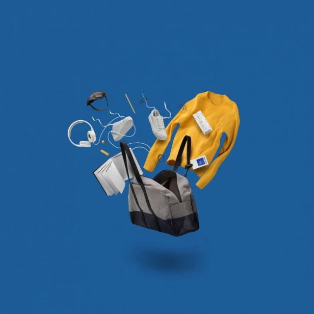 Elevated view of a gray duffel bag surrounded by a yellow sweater, white shoes, headphones, a book, and other objects against a blue backdrop.