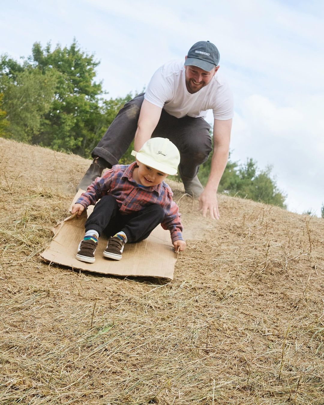 A happy child slides down a grassy hill on a piece of cardboard with help from an adult, enjoying outdoor fun in daylight.