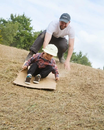 A happy child slides down a grassy hill on a piece of cardboard with help from an adult, enjoying outdoor fun in daylight.