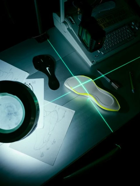 Overhead shot of a shoe sole on a desk with laser beams, design sketches, a magnifying glass, and a computer monitor.