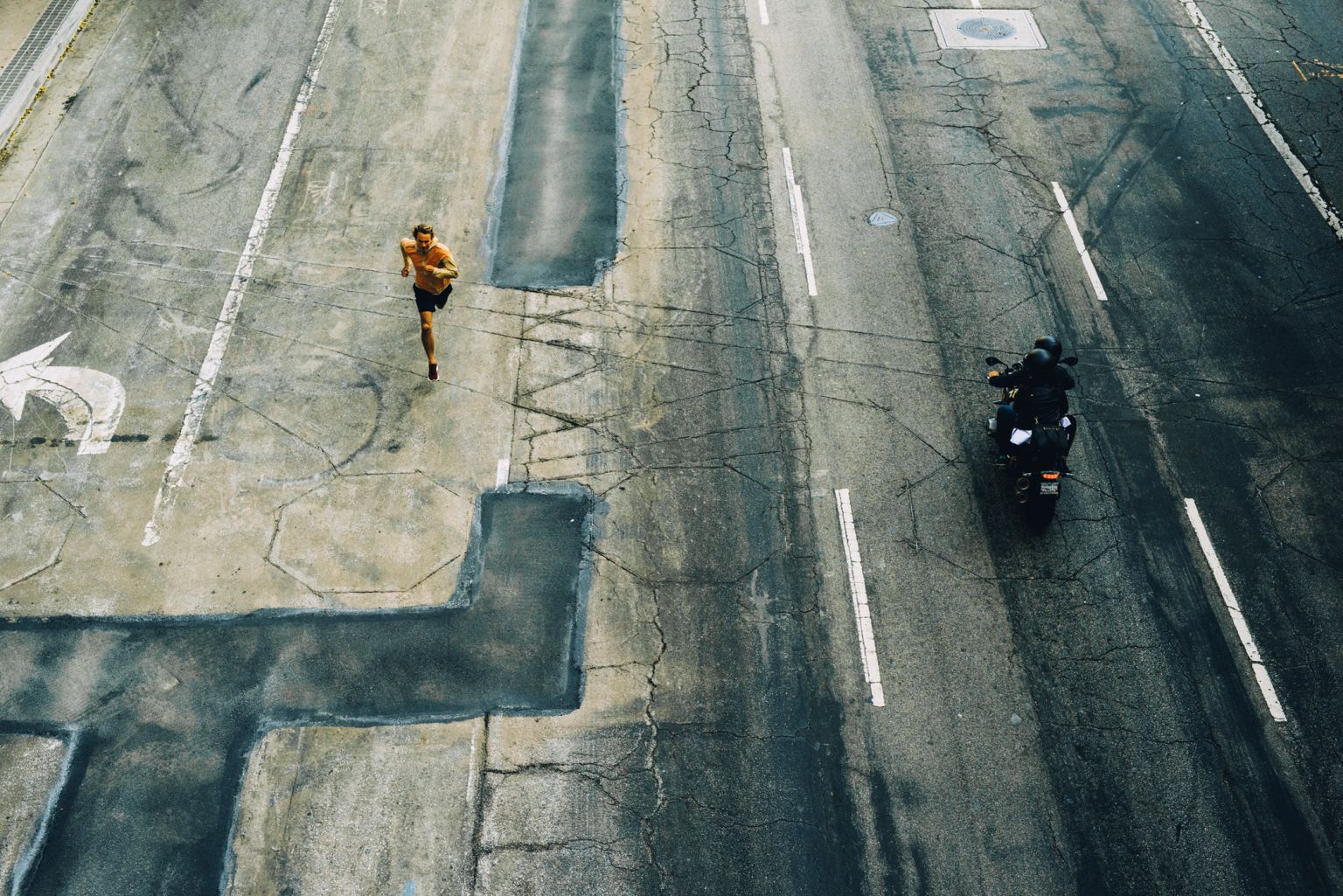 High-angle shot of a man jogging on cracked asphalt next to a motorcycle and rider, showcasing urban activity and contrasting modes of transport.