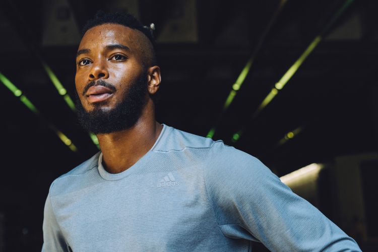 Portrait of a bearded, adult man with dark hair and light blue athletic wear, posed indoors with dark background and soft lighting.
