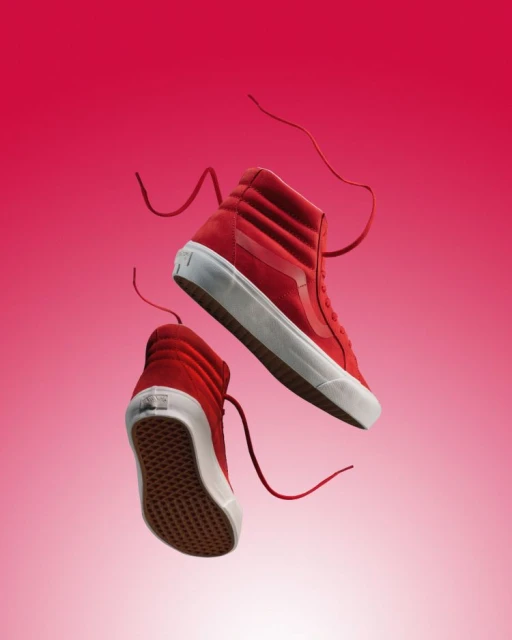 A pair of red Vans high-top sneakers float against a pink gradient background in a studio shot. The shoes are angled showing their soles.