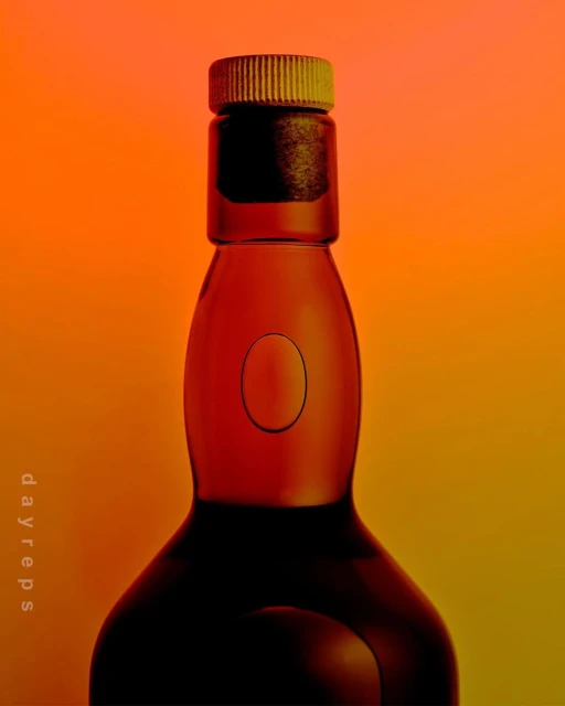 A glass bottle with a ribbed cap stands against a vivid orange and yellow gradient backdrop, its dark silhouette creating contrast.