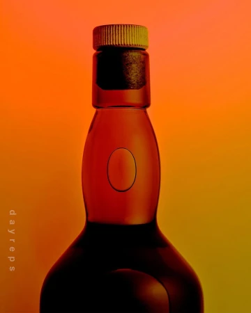 A glass bottle with a ribbed cap stands against a vivid orange and yellow gradient backdrop, its dark silhouette creating contrast.