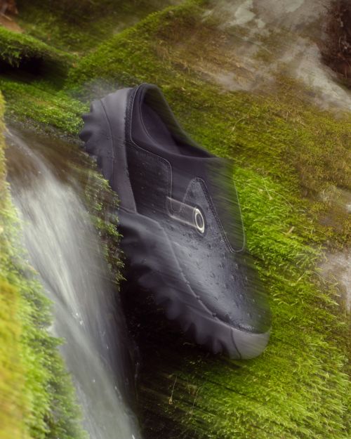 Close-up of a black shoe resting on a mossy surface with water cascading over it in a blurred motion, creating a natural display.