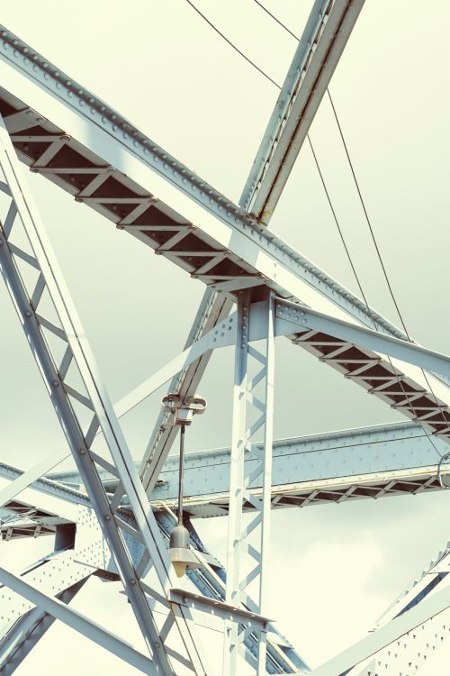 Detailed view of a steel truss bridge structure with an attached street lamp, showcasing complex geometric patterns against a light sky.