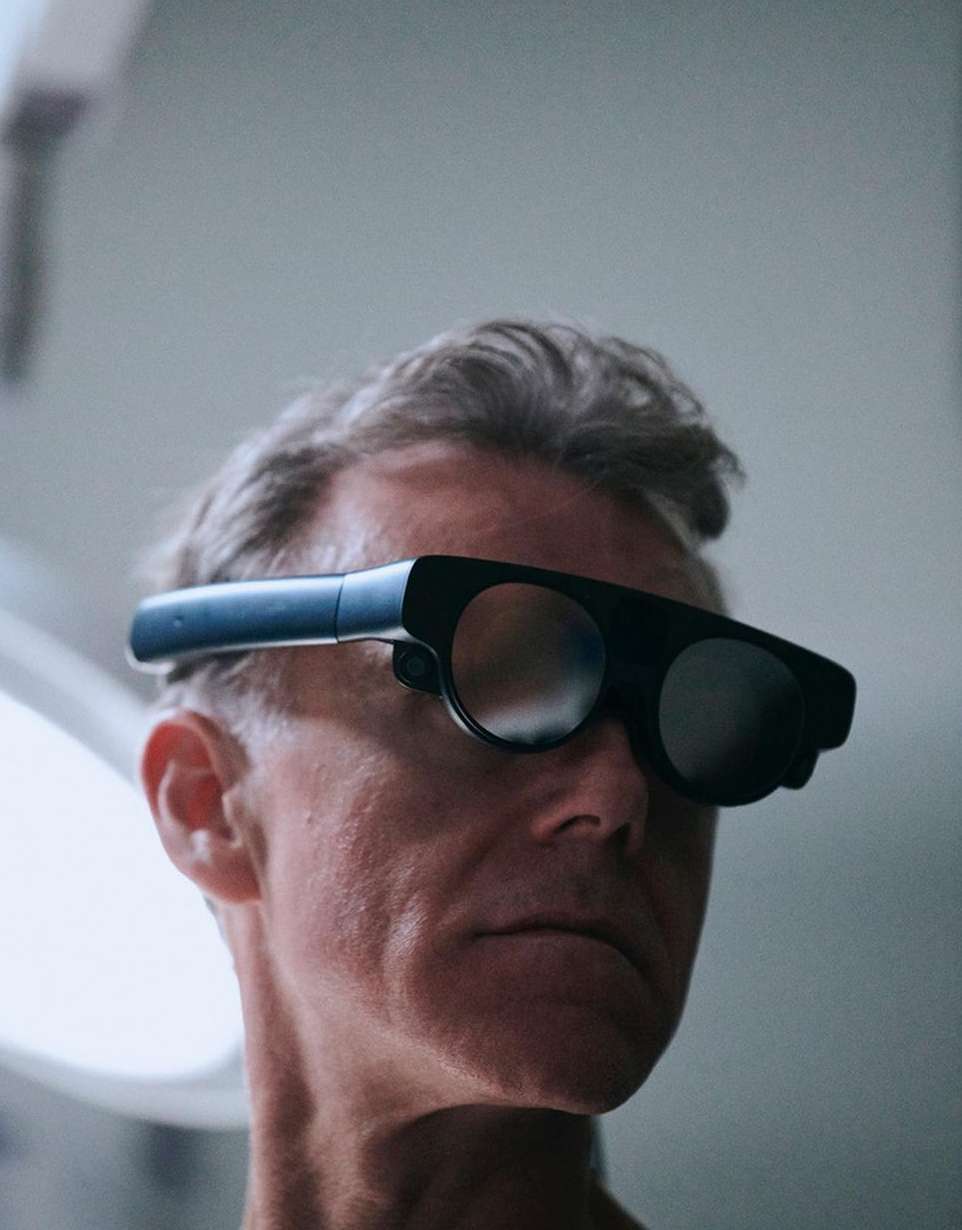 Adult man wearing futuristic augmented reality glasses against a light gray background in a studio or interior setting.