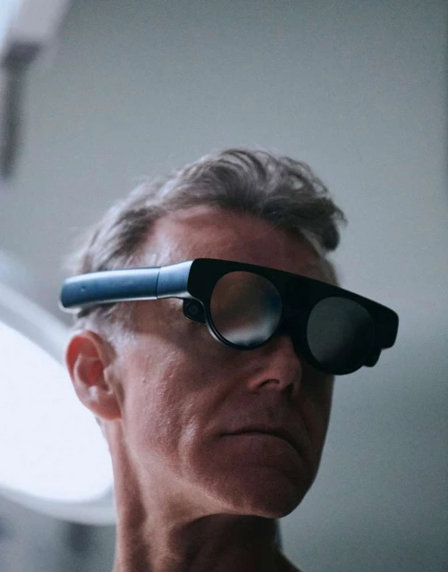 Adult man wearing futuristic augmented reality glasses against a light gray background in a studio or interior setting.