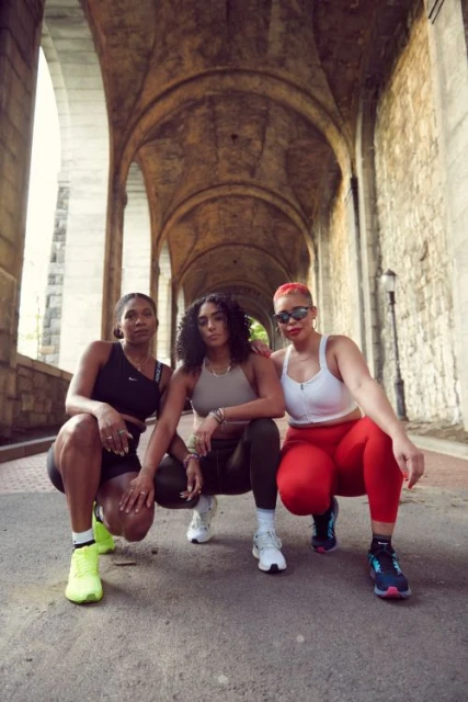 Three female athletes squat side-by-side in a stone archway, wearing workout gear and sneakers, showcasing fitness and outdoor exercise.