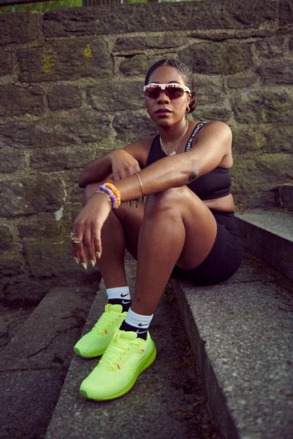 A young woman in athletic wear sits on stone steps wearing sunglasses, a sports bra, bike shorts, neon yellow sneakers, and jewelry.