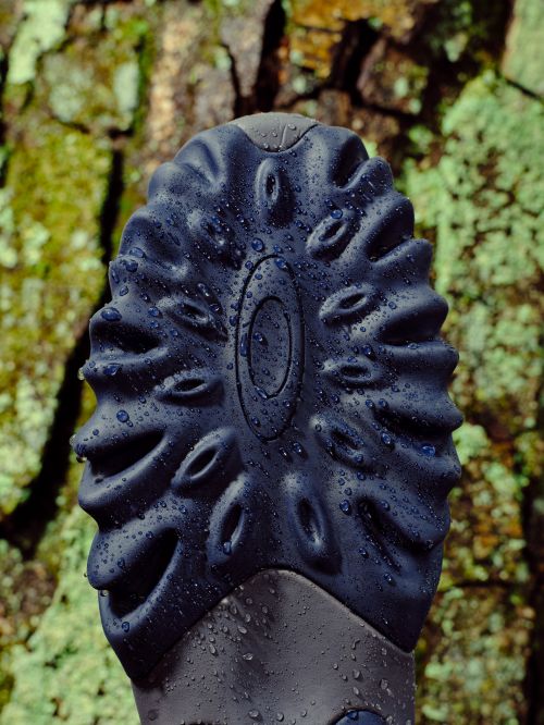 Close-up of a wet, patterned black rubber shoe sole with elliptical openings, set against a background of textured, lichen-covered tree bark.