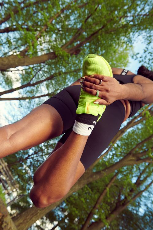 A dark-skinned woman stretches her leg in a green athletic shoe outdoors, showcasing fitness and flexibility against a leafy backdrop.