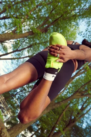 A dark-skinned woman stretches her leg in a green athletic shoe outdoors, showcasing fitness and flexibility against a leafy backdrop.