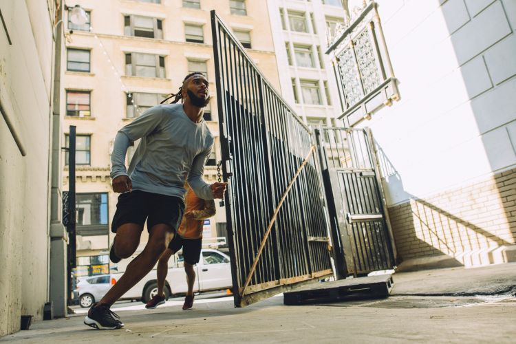 Two athletic adult males are running in an alley. Building, concrete ground, fence, and car in the background under daylight.