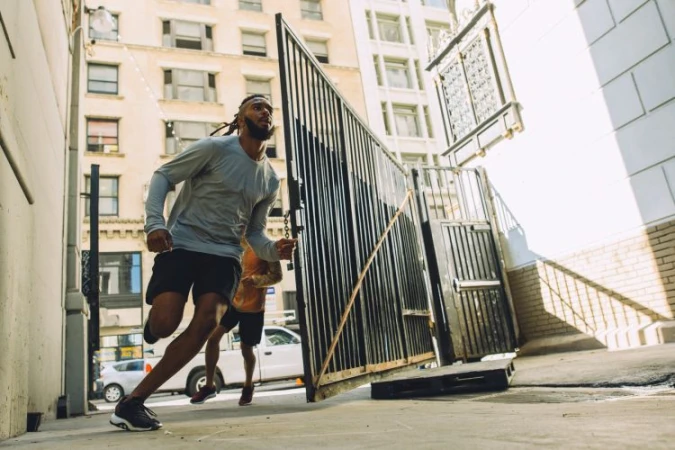 Two athletic adult males are running in an alley. Building, concrete ground, fence, and car in the background under daylight.