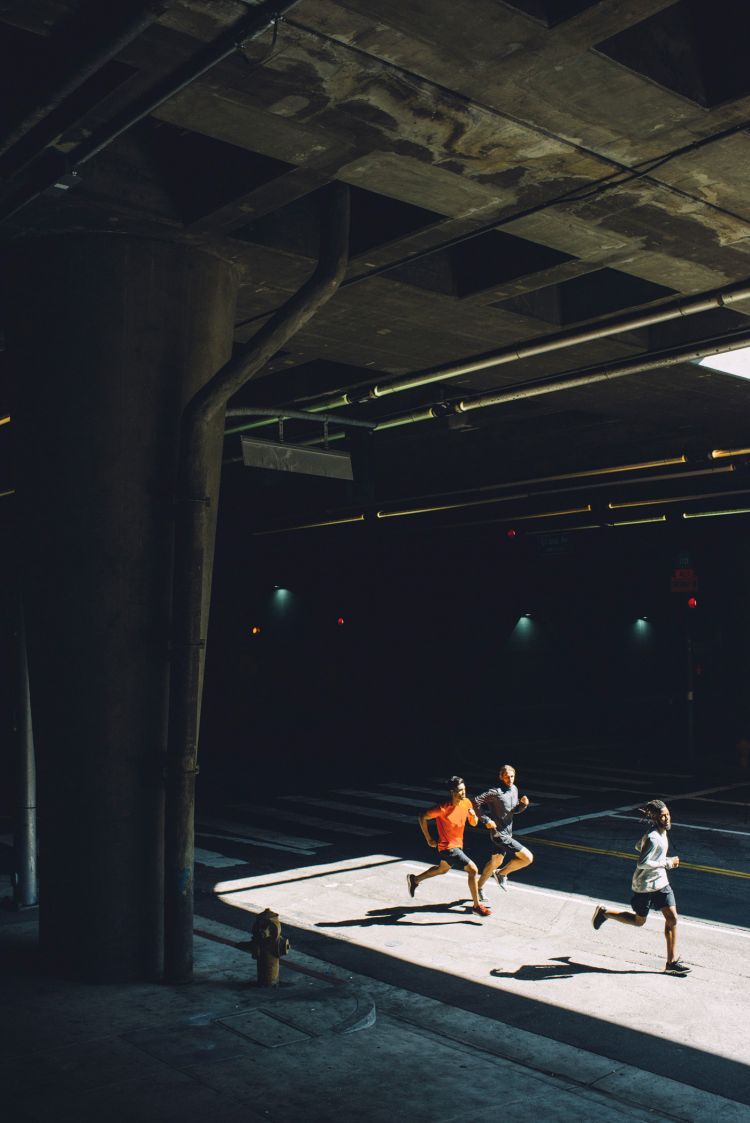 Three men jogging along a concrete sidewalk next to a road, bathed in sunlight streaming from under a large concrete bridge structure.