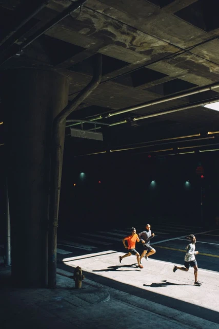 Three men jogging along a concrete sidewalk next to a road, bathed in sunlight streaming from under a large concrete bridge structure.