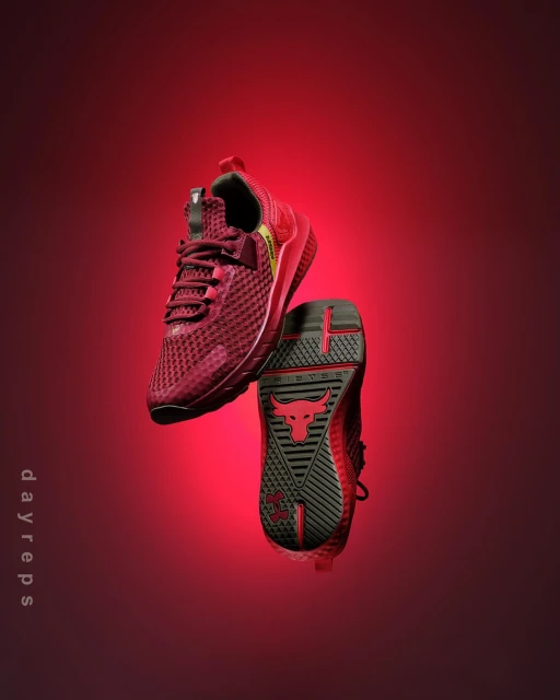 Close-up studio shot of Under Armour Project Rock training shoes in red and black against a vibrant gradient red background.