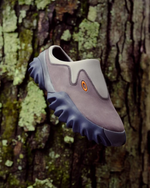 Studio shot of a grey, slip-on Oakley shoe with a rugged sole, showcased against a detailed tree bark backdrop. Focus on footwear.