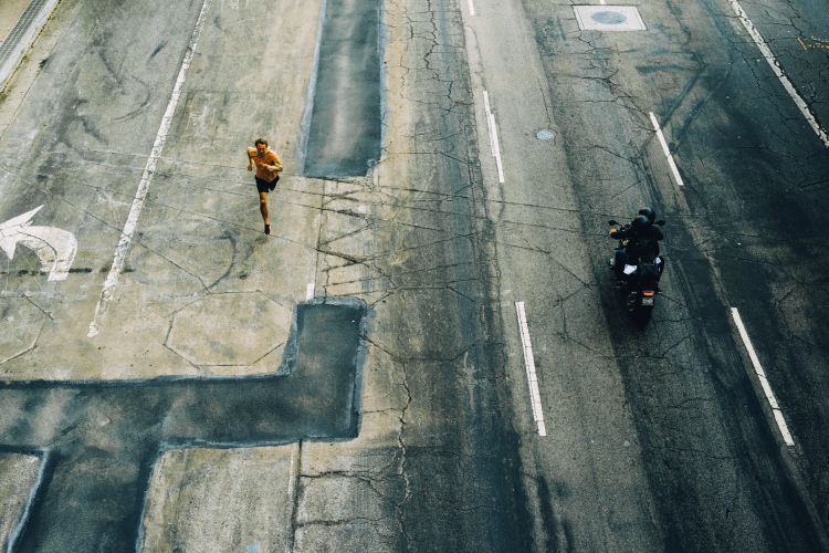 Aerial view of a man running on a cracked road with directional arrows and a motorcyclist in the opposite lane.
