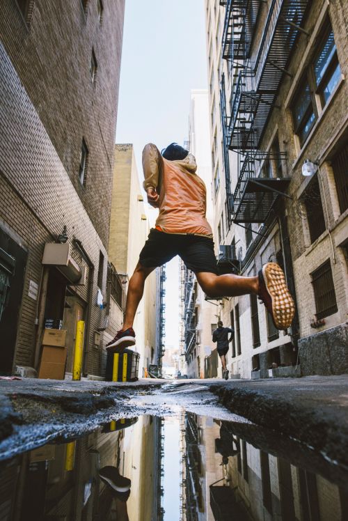 Person leaps over a puddle in an urban alley, with buildings and another runner visible in the background, captured at a low angle.