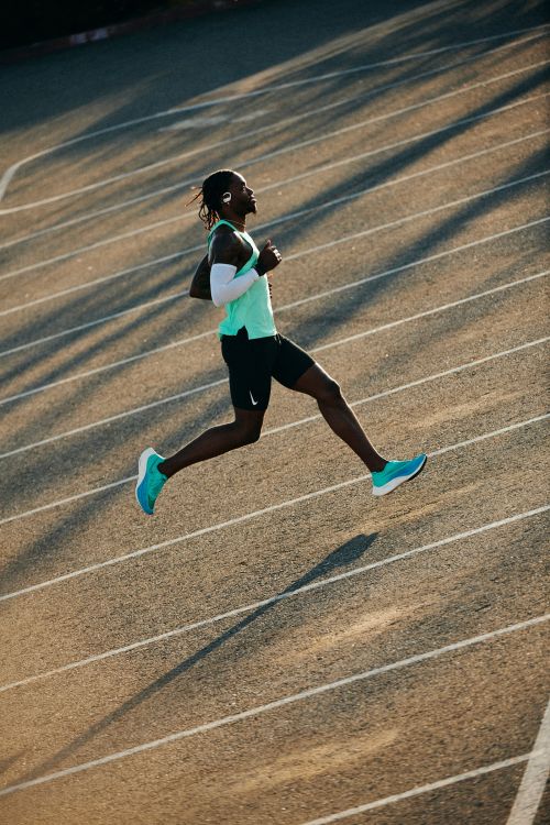 An African-American man runs at speed on a running track wearing athletic gear and wireless headphones in the sunlight. 