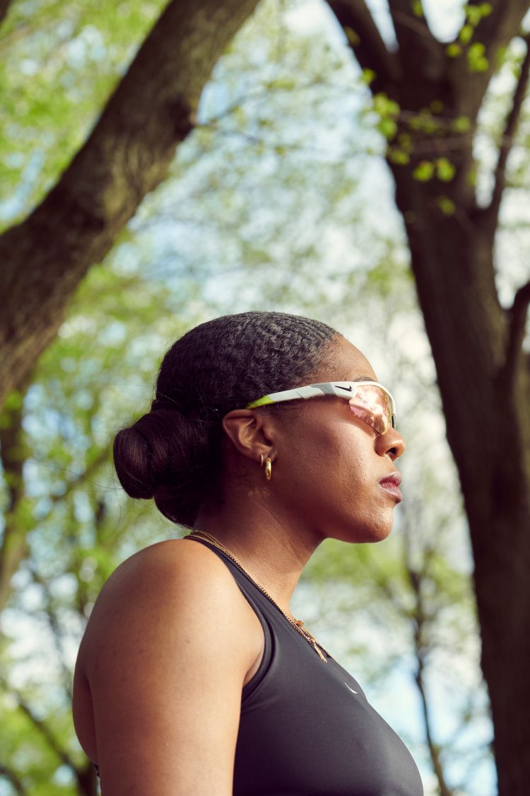 Black woman wearing sports sunglasses and a black athletic top outdoors against a backdrop of trees